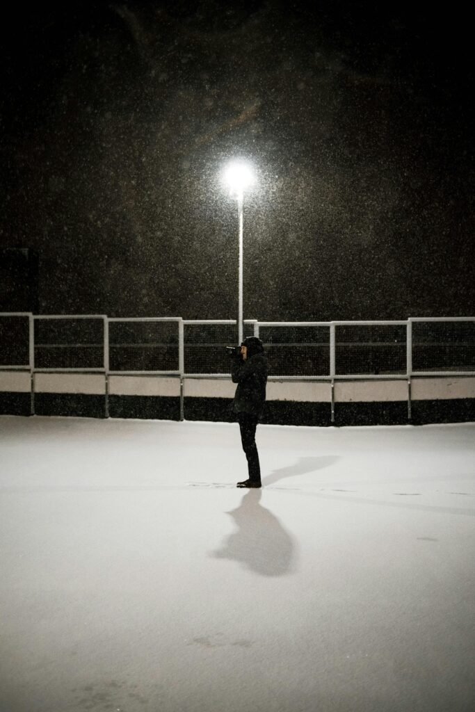A solitary person stands in the snow, illuminated by a street lamp, creating a serene winter scene.