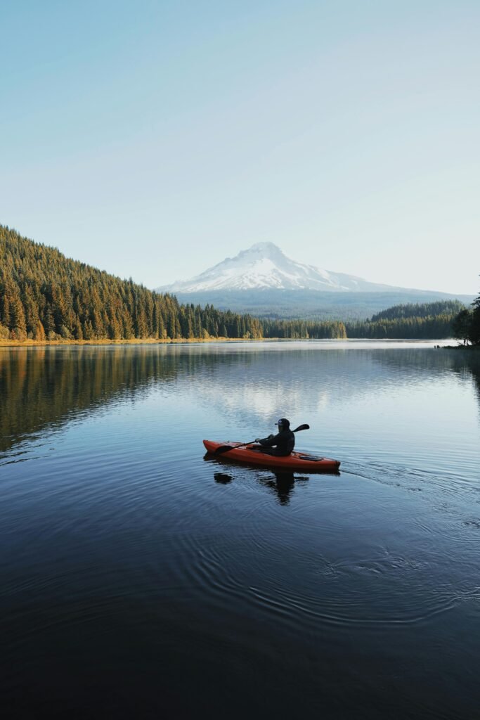 A kayaker paddles on a placid lake surrounded by forest with a snow-capped mountain in the background.