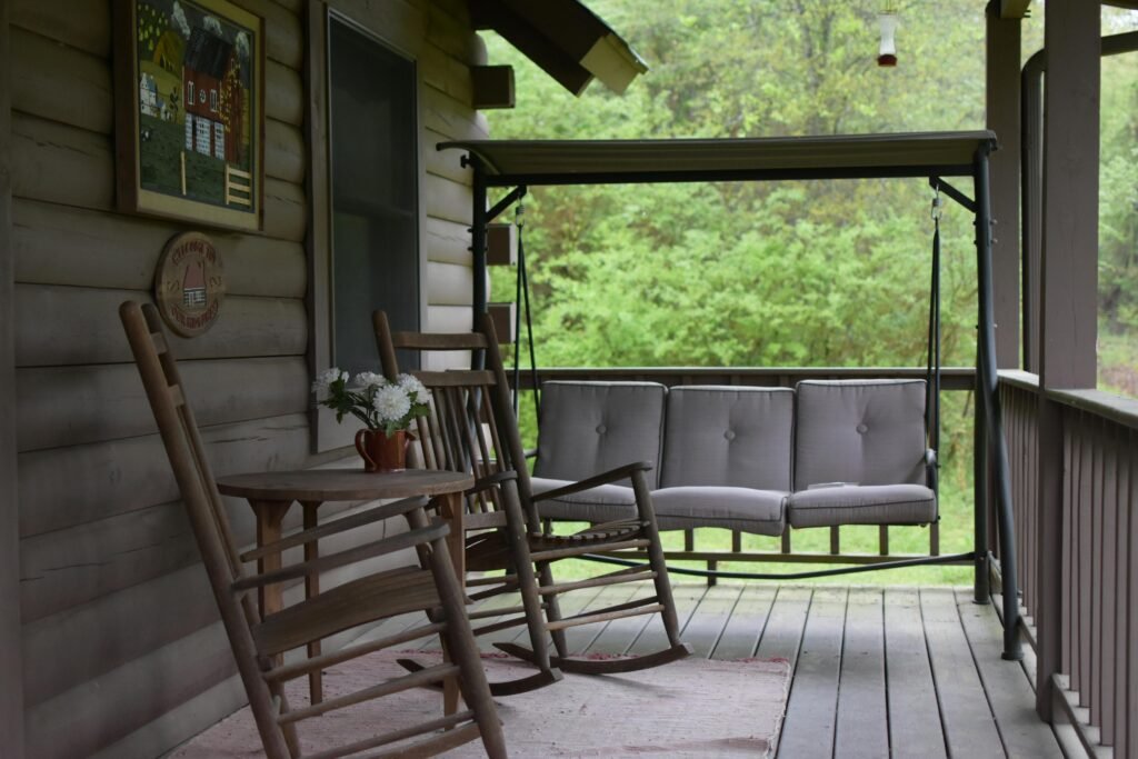 Cozy rustic porch with wooden rocking chairs and a swing, surrounded by lush greenery.